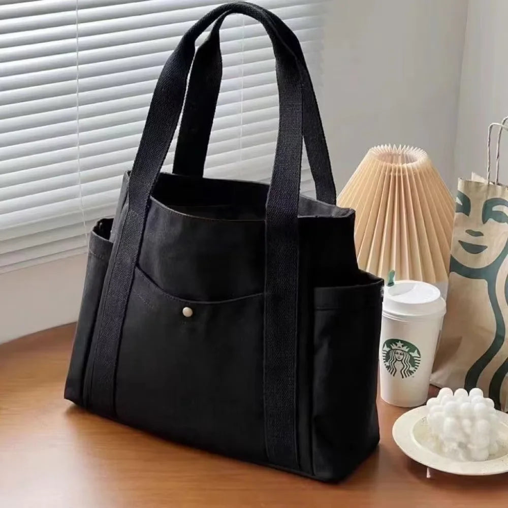 A black canvas tote bag with a zipper closure is displayed on a wooden surface next to a paper bag.