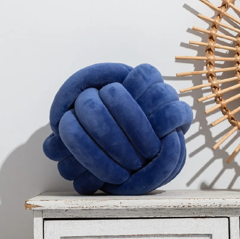 A deep blue velvet knitted throw pillow resting on a white wooden surface next to a woven sunburst decoration.
