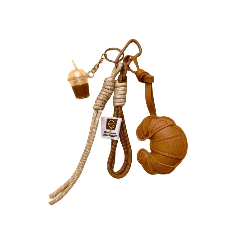 A braided leather croissant keychain featuring a bag pendant and a small glass charm.