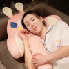 A person is resting their head on an adorable pink plush cat and dog backrest cushion while relaxing on a sofa.