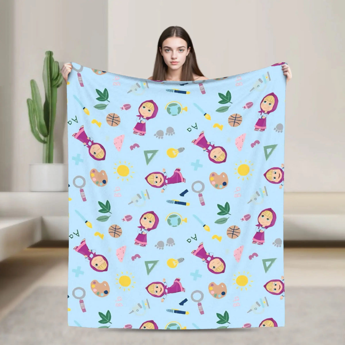 A woman holds up a light blue flannel throw blanket featuring cartoon designs of Masha and Bear.