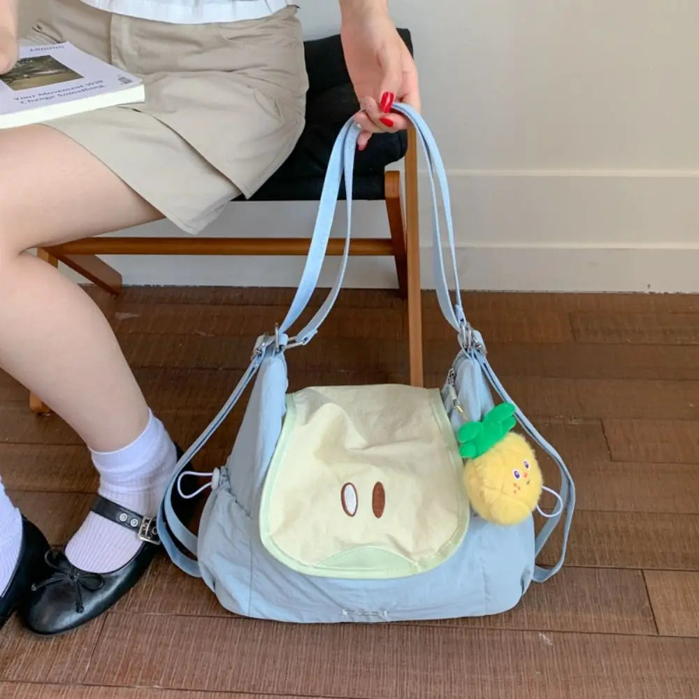 A light blue nylon messenger bag with a yellow fruit charm is being held by a person sitting on the floor.