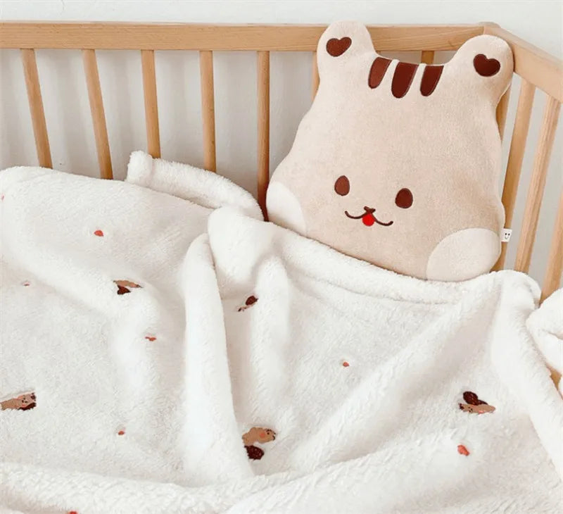 A soft, cream-colored coral fleece baby blanket with bear embroidery draped over a wooden crib rail.