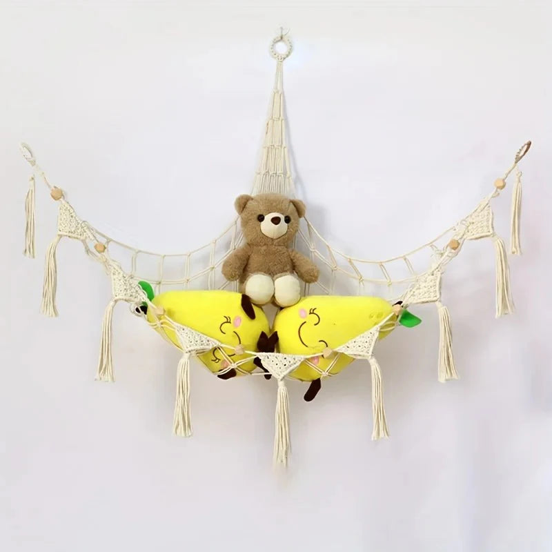A bohemian plush toy storage hammock made of macrame netting with hanging tassels, holding a brown teddy bear and two yellow 