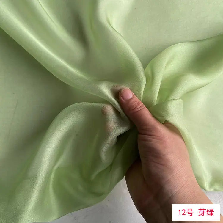 A hand is folding a piece of light green silk chiffon fabric to demonstrate its drape and texture.