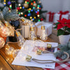 String lights with gold-toned bulbs are arranged on a wooden table next to a Christmas tree and wrapped gifts.