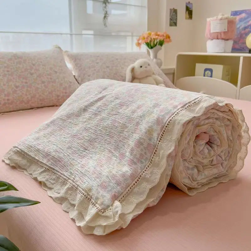 A rolled floral washed cotton gauze summer quilt with delicate lace trim resting on a pink surface.