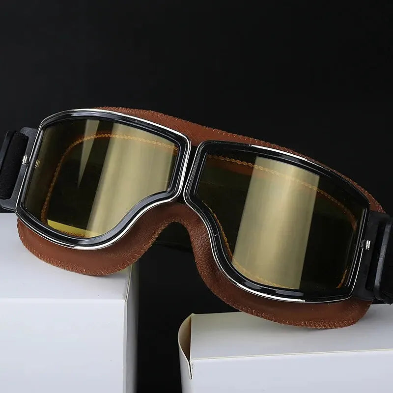 A pair of brown retro foldable motorcycle goggles with gold tinted lenses resting on a white box.