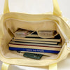A pale yellow shoulder bag with a lace trim, featuring a clear pocket holding books and a smartphone.