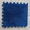A rectangular blue plush interlocking floor tile resting on a light-colored, perforated surface.