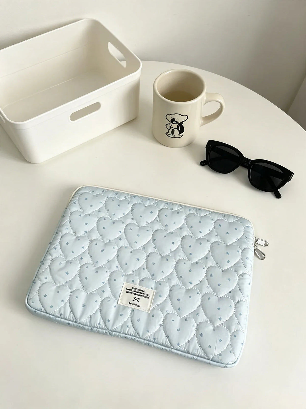 A light blue quilted laptop sleeve with a soft lining resting on a white table next to a mug and sunglasses.