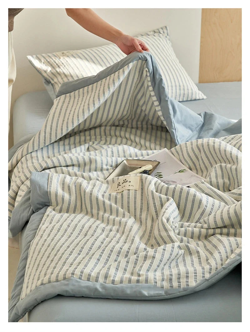 A light blue and white striped breathable summer quilt draped over a bed, with a small wooden item resting on top.