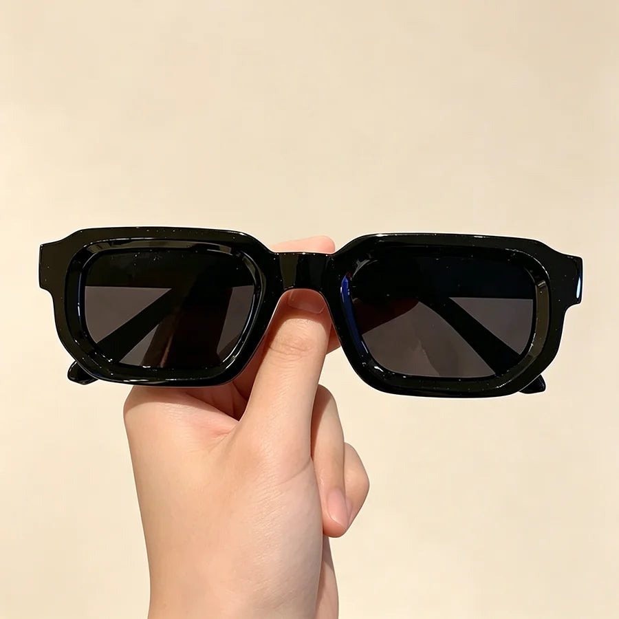 A hand holds a pair of black vintage rectangle sunglasses against a light background.