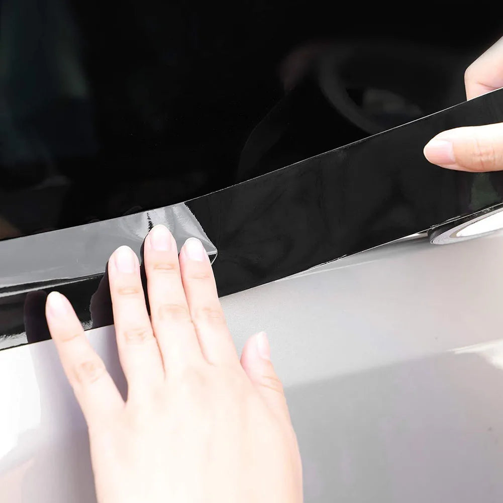 A hand is applying a high-gloss black vinyl wrap strip to the edge of a car body.