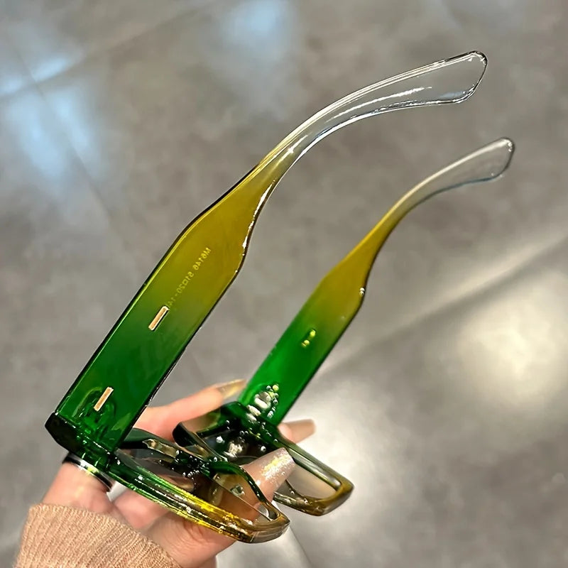 A pair of gradient square sunglasses with green and yellow lenses held in a hand.