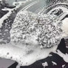 A white microfiber car wash mitt is shown resting on a dark surface covered in soapy water and bubbles.