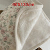 A white floral muslin baby blanket with a textured, fluffy trim is shown folded.
