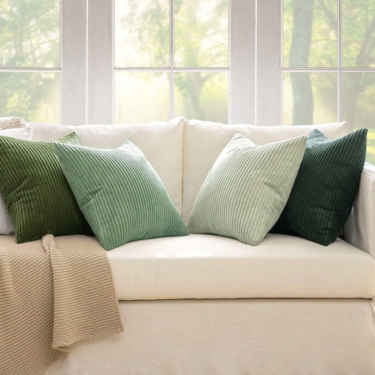 A collection of olive green and sage colored corduroy throw pillow covers arranged on a light beige sofa near a window.
