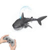 A gray shark toy is shown spraying water, with a hand holding a remote control nearby.
