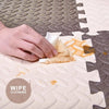 A hand is using a white wipe to clean a section of a tan interlocking foam play mat.