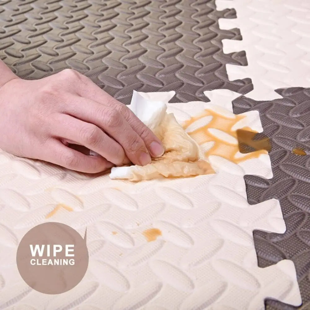 A hand is using a white wipe to clean a section of a tan interlocking foam play mat.