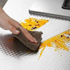 A hand is using a brown scrubbing pad to clean spilled yellow oil on a white surface.