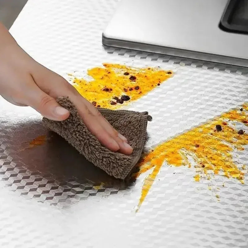 A hand is using a brown scrubbing pad to clean spilled yellow oil on a white surface.