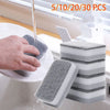 A hand is using a gray scouring pad to clean a plate under running water, with other stacked sponges visible.