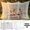 A large white mesh mosquito net canopy is set up outdoors, draped over a wooden table where a family is having a meal.