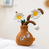 A brown plush doll shaped like a plant with white flowers and green leaves resting on a white surface.