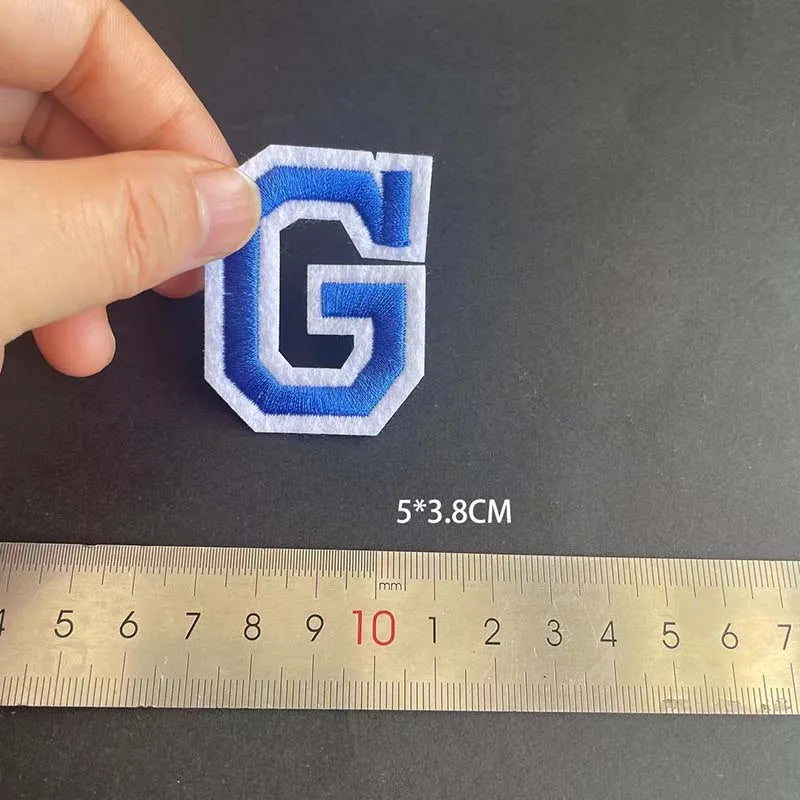 A hand holds a blue embroidered letter G patch, measured against a ruler.