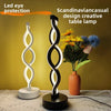 A black and white, seaweed-shaped desk lamp with a soft glow sits next to a vase of yellow flowers on a light surface.