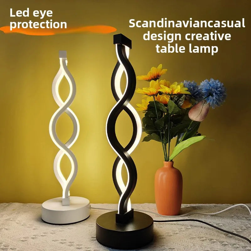 A black and white, seaweed-shaped desk lamp with a soft glow sits next to a vase of yellow flowers on a light surface.