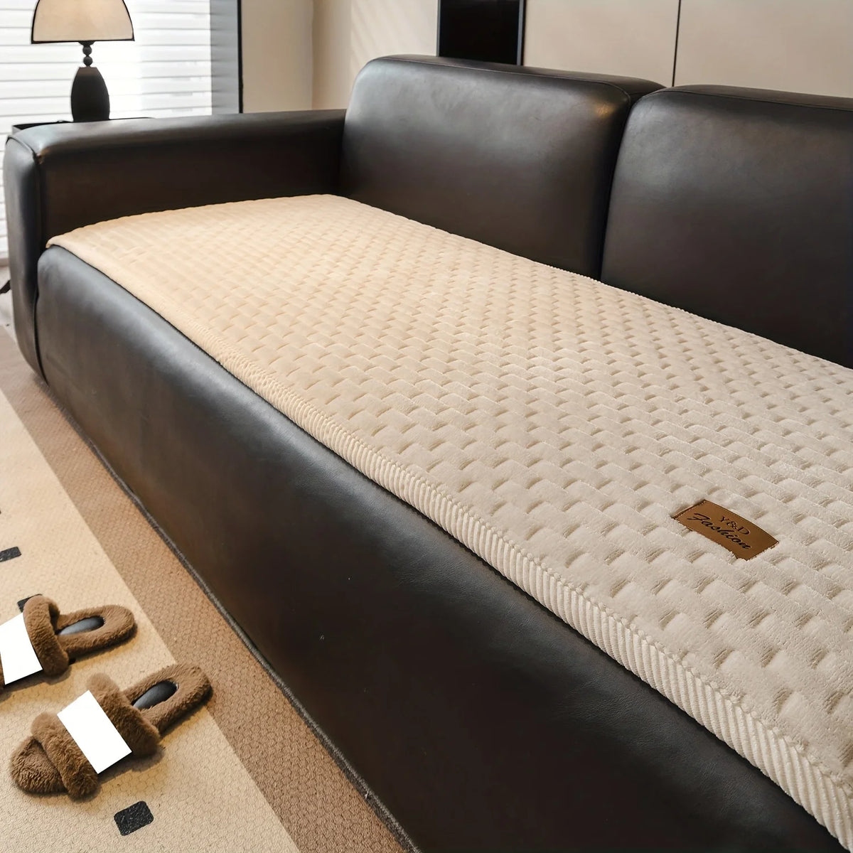 A dark brown leather sofa with a beige quilted plush cover and non-slip backing on a light carpet.