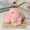 A pink fluffy angel bunny keychain with a pom pom tail is displayed on a wooden tray.