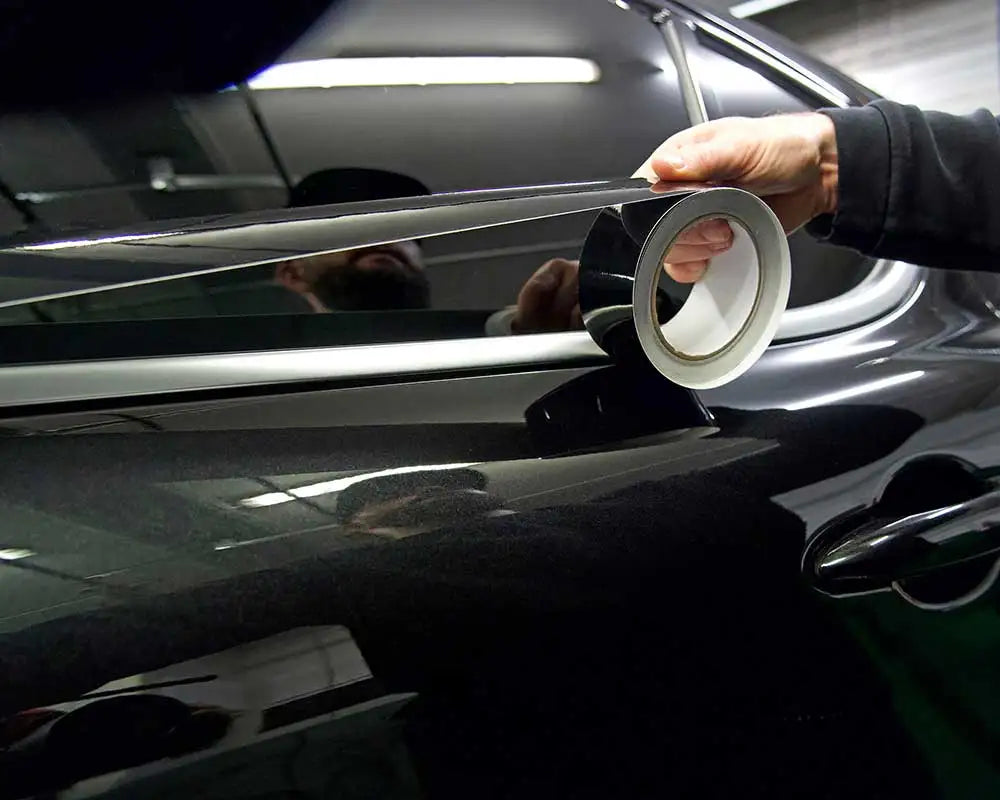 A hand is applying a glossy black vinyl wrap strip to the trim of a dark car.