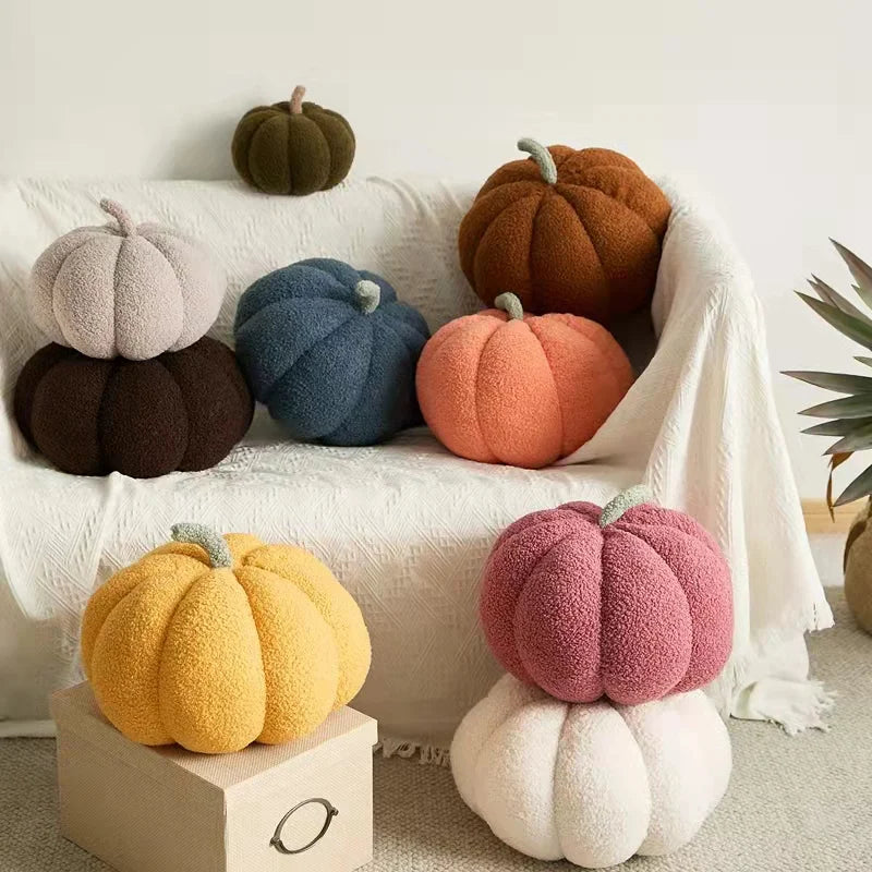 A collection of soft, round plush pumpkins in various earthy tones like orange, brown, and gray arranged on a white surface.