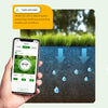 A hand holds a smartphone displaying a smart sprinkler controller interface, with water droplets falling onto green grass.