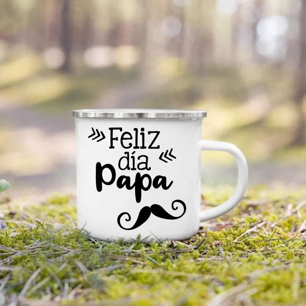 A white enamel camping mug with black text that reads Feliz día papa and a mustache design, placed on green grass.
