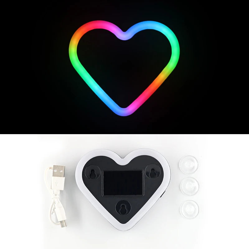 A glowing heart-shaped LED light ring in a rainbow gradient, next to a black heart-shaped device and some clear lenses.