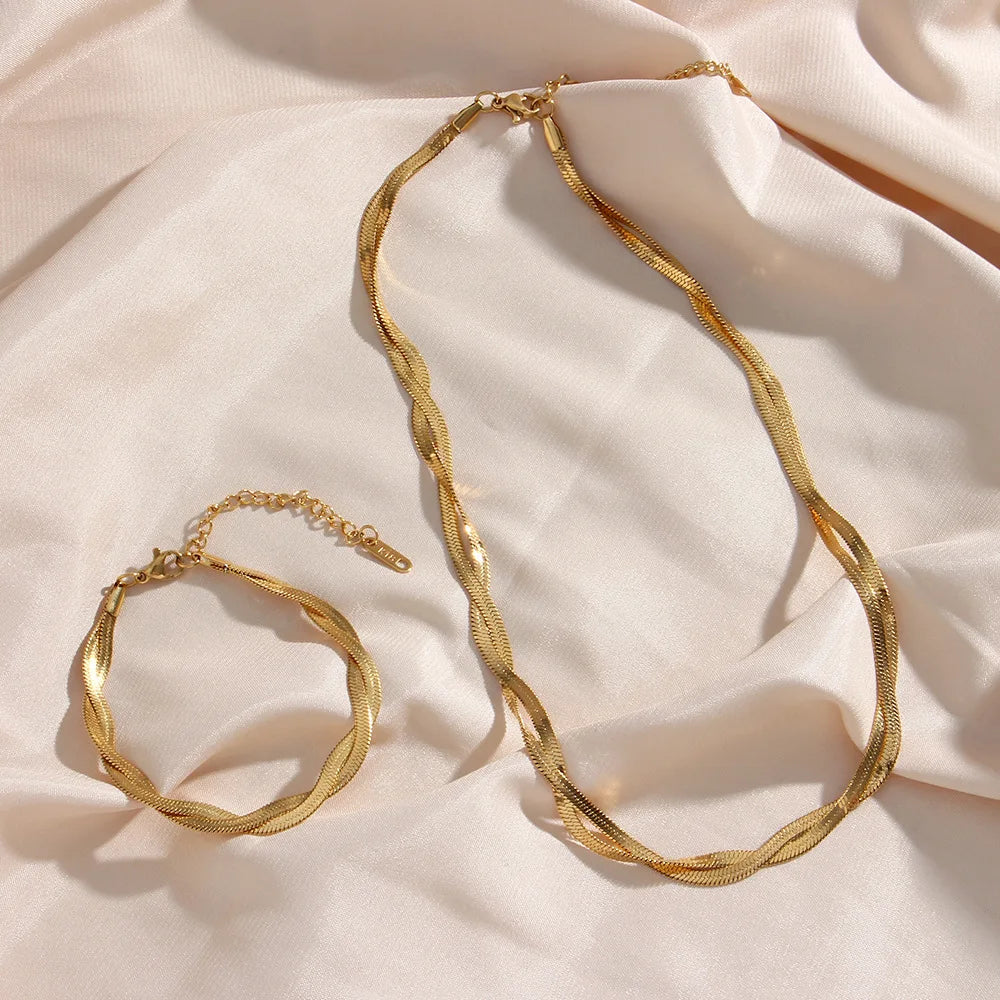 A set of gold-plated herringbone chain necklace and bracelet displayed against a soft beige fabric background.