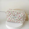 A floral quilted makeup bag with a white background and pink, red, and green flowers is displayed on a round white stand.