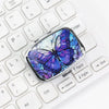 A portable metal pill organizer and jewelry case featuring a vibrant purple and blue butterfly design placed on a white lapto