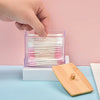 A hand is inserting a cotton swab into a clear storage dispenser filled with pink and white swabs, resting on a wooden surfac