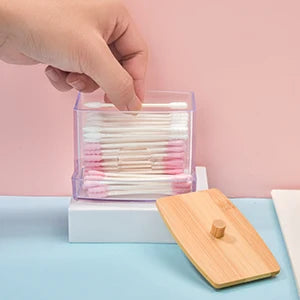 A hand is inserting a cotton swab into a clear storage dispenser filled with pink and white swabs, resting on a wooden surfac