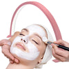 Hands apply a white facial mask to a person's face under a pink LED arc lamp.
