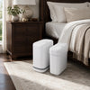 A white, rectangular trash can is placed on a light rug next to a wooden bed frame.