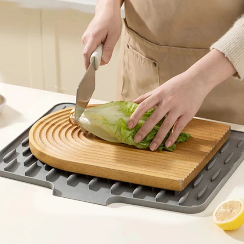 A hand is using a knife to cut lettuce on a wooden cutting board placed on a silicone sink drain mat.