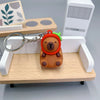 An adorable capybara keychain charm with an orange body and green ear, attached to a silver chain, resting on a wooden shelf.
