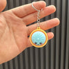 Eine Hand hält einen kleinen gelb-blauen dekorativen Dumpling-Anhänger mit einem silbernen Ring.